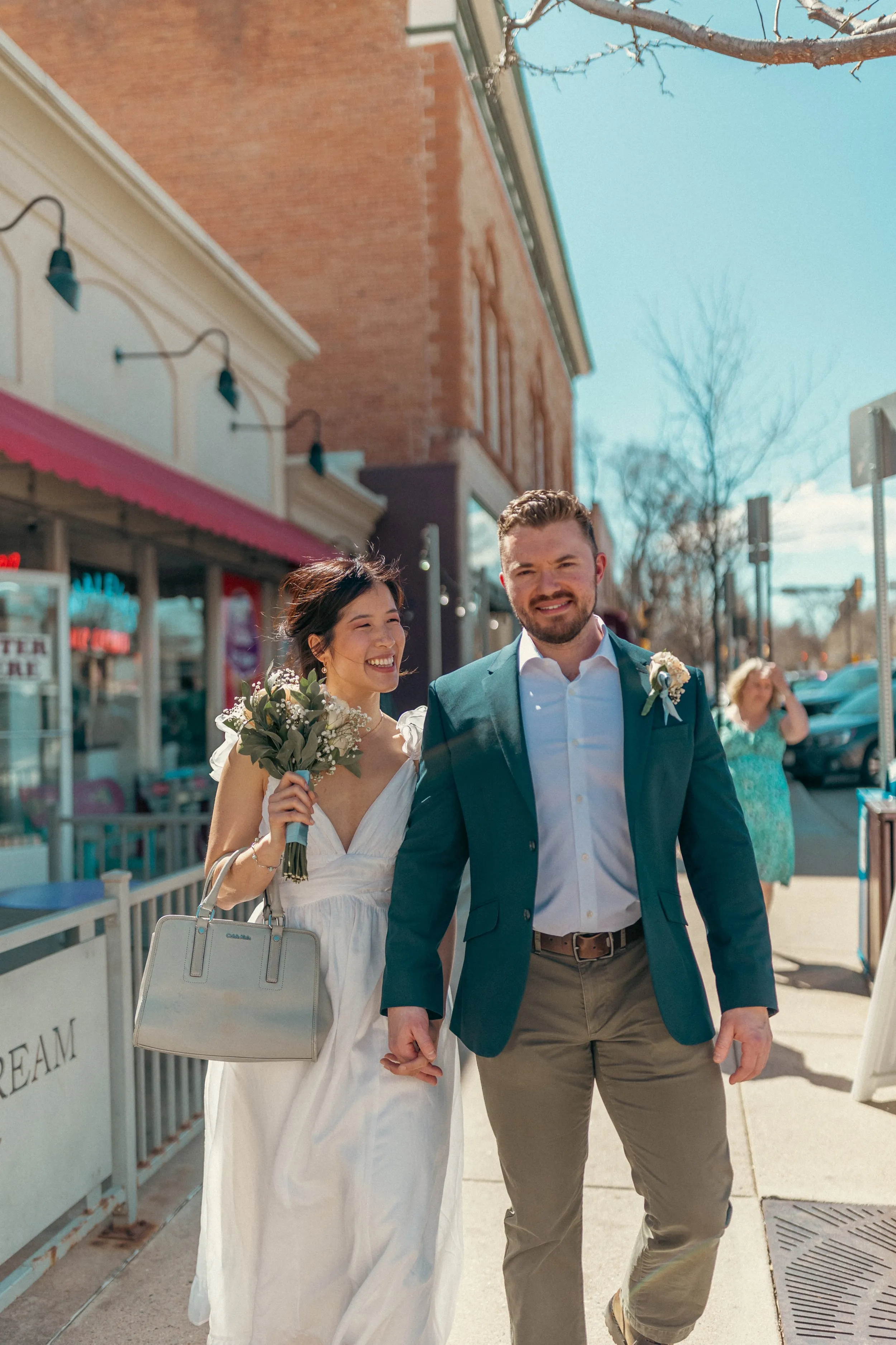 Newlyweds walking a sunlit street hand in hand, the bride holding her bouquet