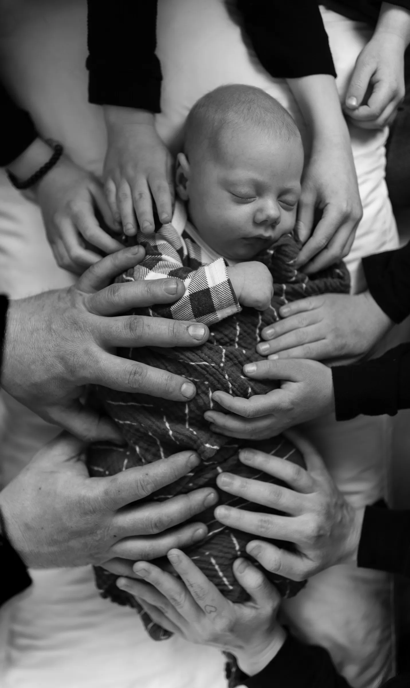 Many hands cradling a swaddled newborn, in black and white